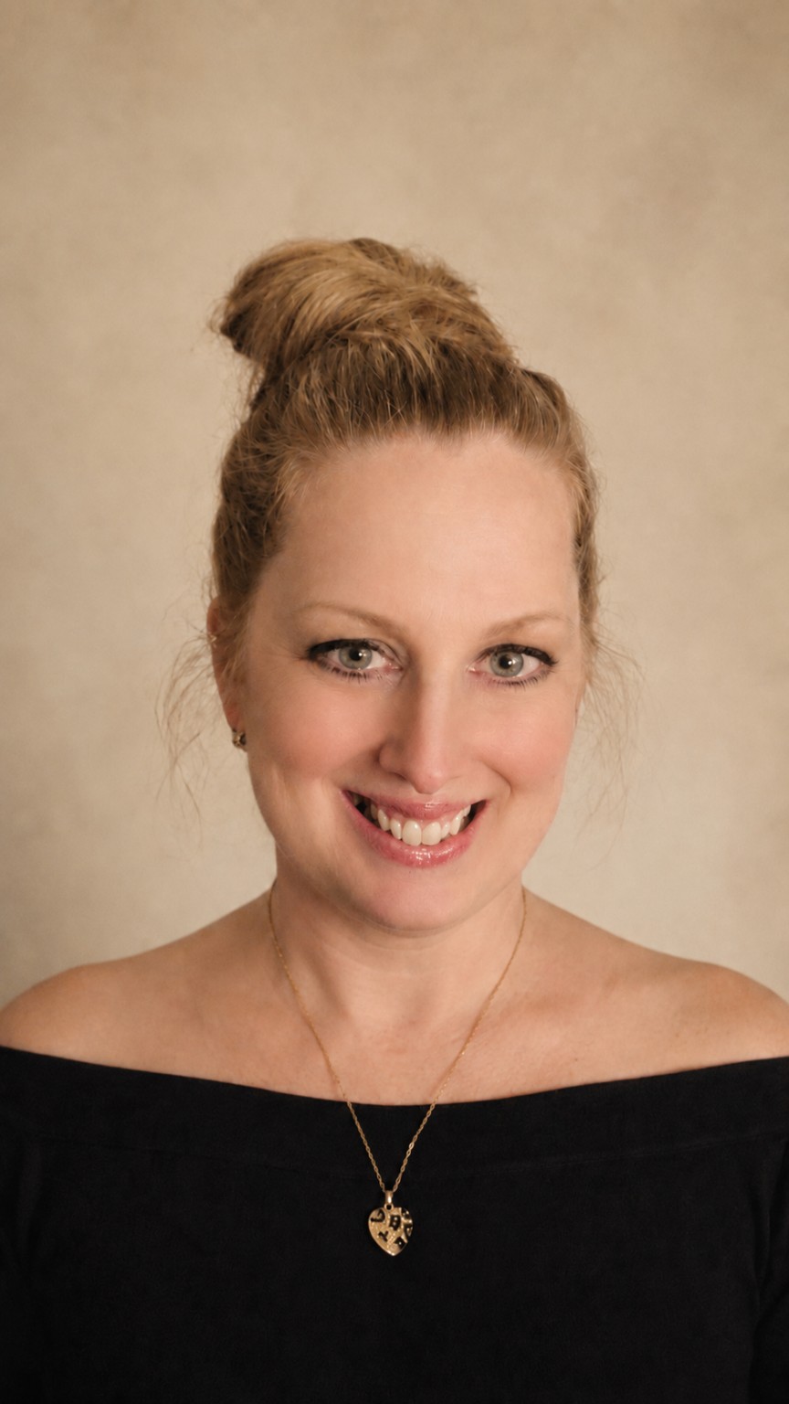 Portrait of Diane Cassi smiling, her hair in a bun, wearing a black off-the-shoulder top and a gold heart pendant necklace against a neutral beige background